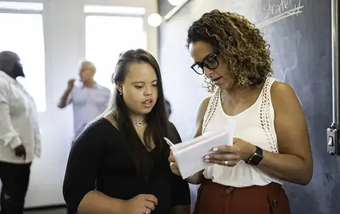 Two women standing in front of a blackboard, engaged in discussion or teaching, with educational materials visible.