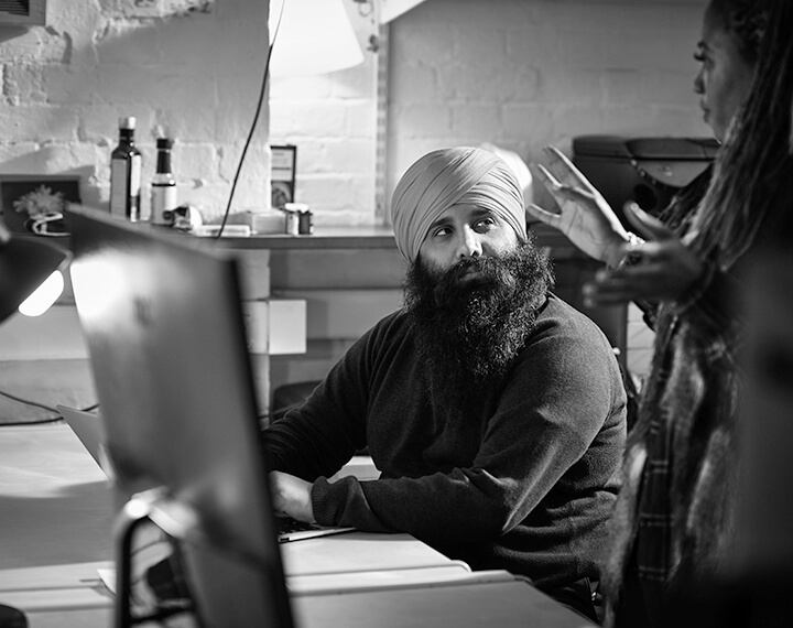A bearded man wearing a turban is seated at a desk, engaged in work or contemplation.