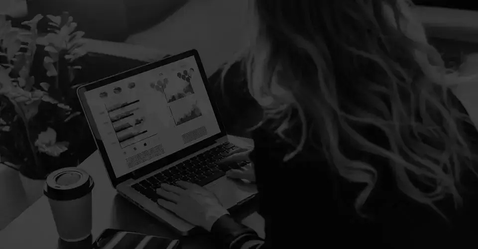 A woman seated at a desk, focused on her laptop, surrounded by a tidy workspace and soft lighting.