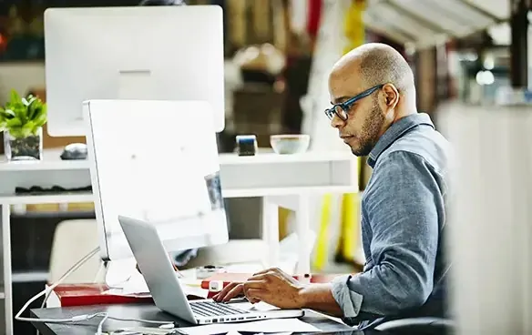 A man wearing glasses is focused on his laptop, engaged in work at a desk.