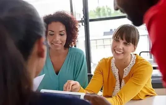 A diverse group of individuals gathered around a table, engaged with a tablet in a collaborative discussion.